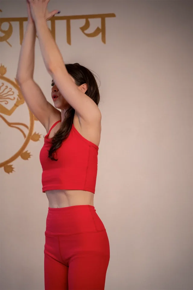Rachael standing with her arms raised overhead, in front of a wall painted with Sanskrit lettering.